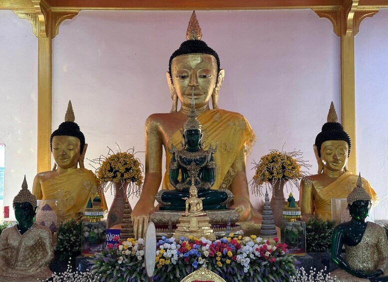 A large Buddha statue flanked by two smaller statues, surrounded by flowers and traditional Thai architecture. 2
