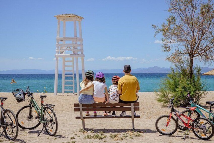 A family sitting on a bench at a beach with bicycles and a lifeguard tower in the background. 1