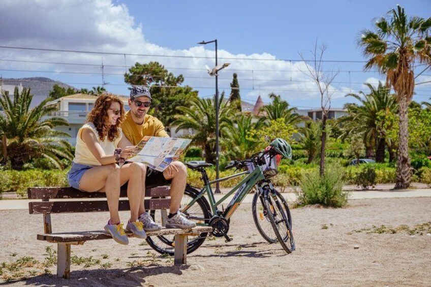 Two people sitting on a bench with a bicycle leaning against it, surrounded by palm trees and a clear sky. 5