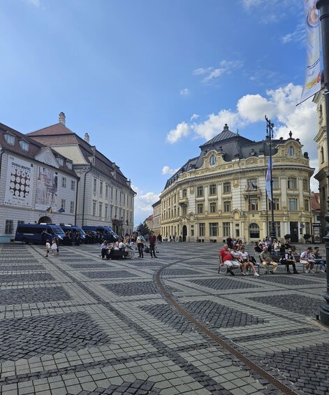 A cobblestone square with historic buildings, a tram track, and people sitting on benches. 3