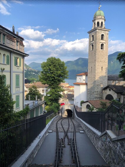 A railway track leading to a tower with a green dome, surrounded by buildings and a mountainous backdrop. 1
