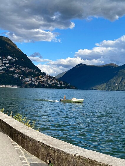 A calm lake with a small boat carrying a person, surrounded by mountains and a town. 4