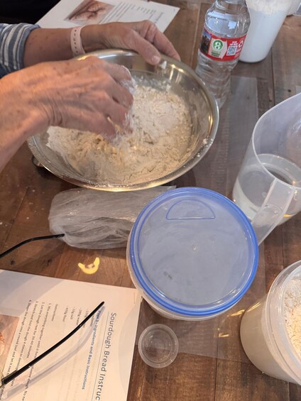 Hands mixing flour in a bowl, with a recipe book, plastic wrap, and a water bottle on a wooden table. 2