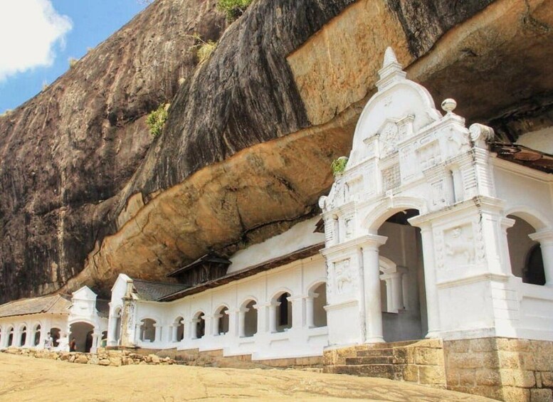 A white temple with arches and columns, set against a large rock formation. 2