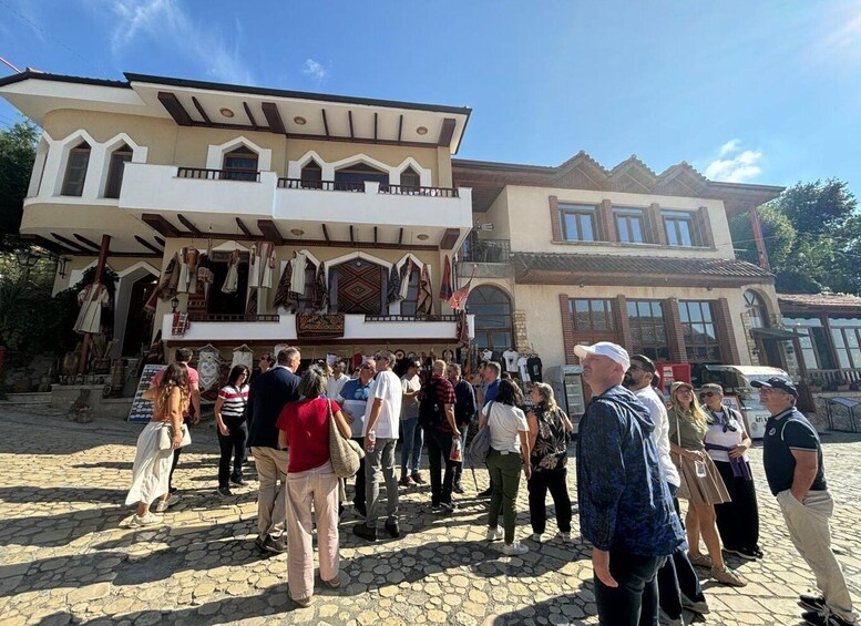 A cobblestone square with a crowd of people, a building with a balcony, and a clear blue sky. 5