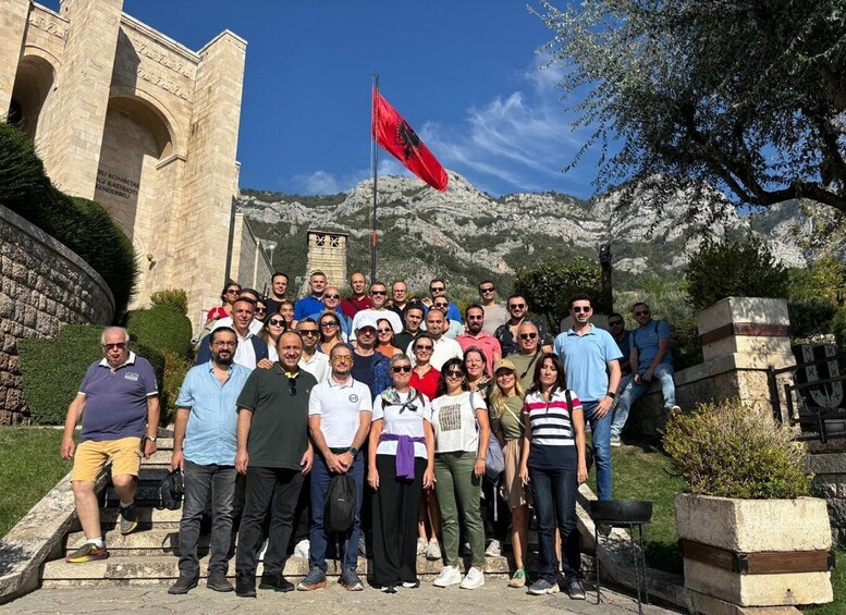 A group of people standing in front of a stone archway with a red flag, with mountains in the background. 2