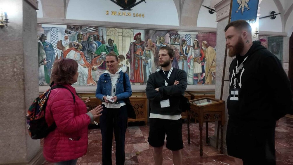 A group of people standing in front of a historical mural with the inscription '2 MARS 1444'. 3