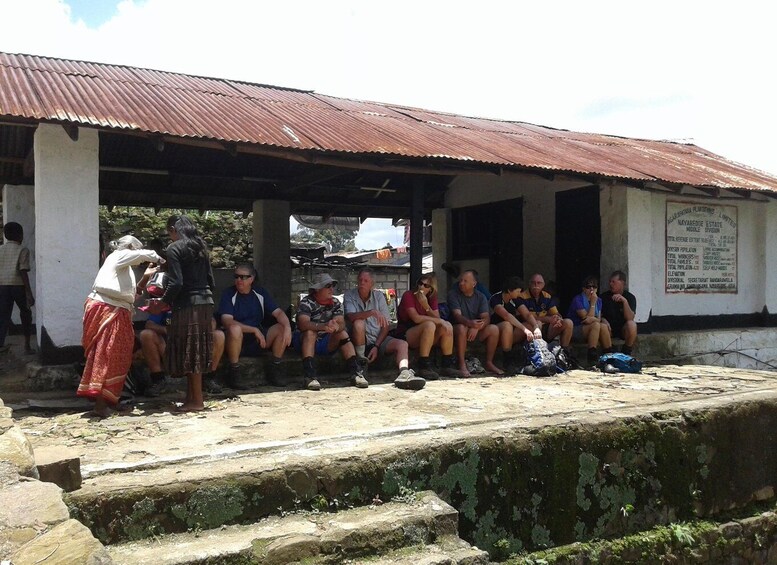 A group of people seated under a corrugated metal roof, with a stone wall and a signboard in the background. 4