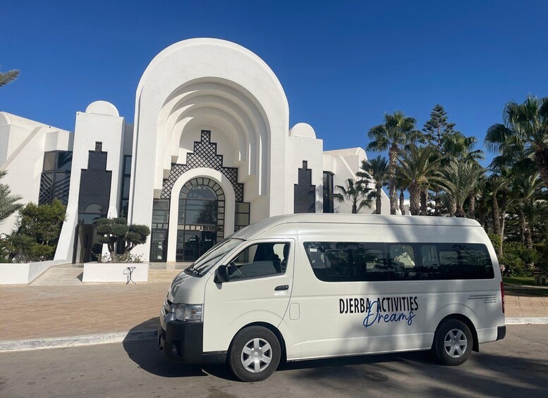 A white van parked in front of a modern building with a large arched entrance and palm trees in the background. 1