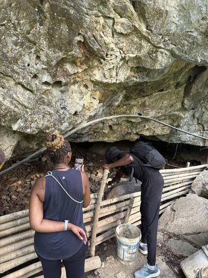 Two people near a cave entrance, one taking a photo, and another inspecting a wooden structure. 4