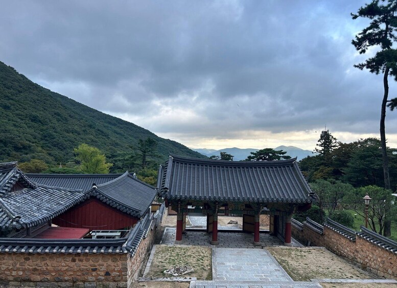 Traditional Korean architecture with tiled roofs and stone walls, set against a backdrop of lush green hills and a cloudy sky. 4