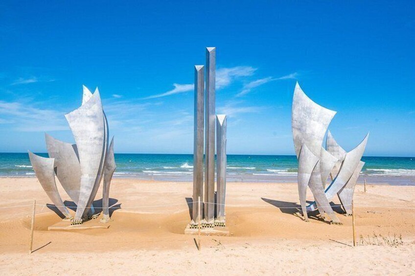 Three metallic sculptures on a sandy beach with the ocean in the background. 3