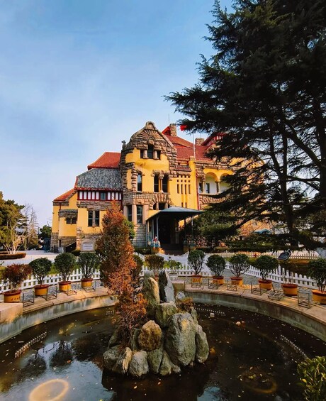 A historic building with a yellow facade, red roof, and a pond with rocks in the foreground. 1