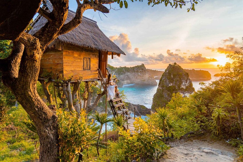 A wooden house on stilts with a thatched roof, overlooking a coastal landscape with cliffs and a sunset. 1