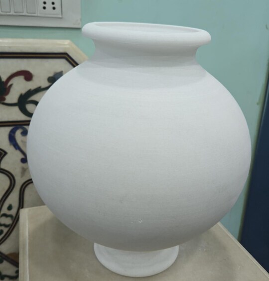 A white ceramic pot with a rounded body and a flared rim, placed on a beige surface against a teal wall with a decorative tile pattern. 2