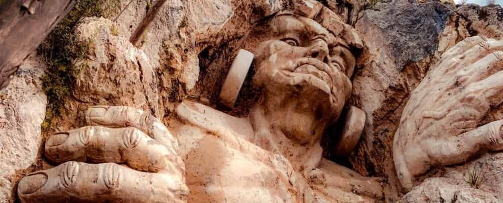 A large stone sculpture of a human figure with hands extended, set in a rocky terrain. 1