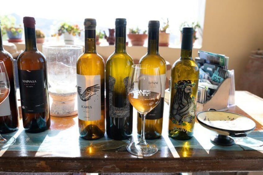 A selection of wine bottles and glasses on a table, with a clear sky and potted plants in the background. 1