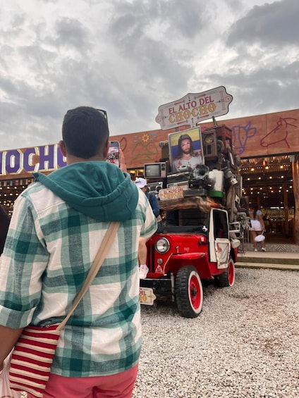 A man in a green and white checkered shirt and red shorts, standing in front of a vintage red car with a sign that reads 'EL ALTO DEL CHOCOLAT'. 5