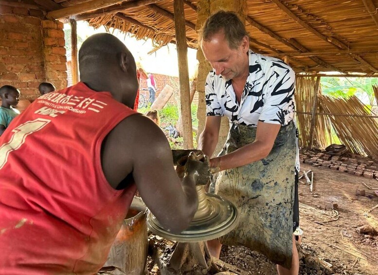 Two people engaged in pottery making, with a thatched roof structure and bricks in the background. 2