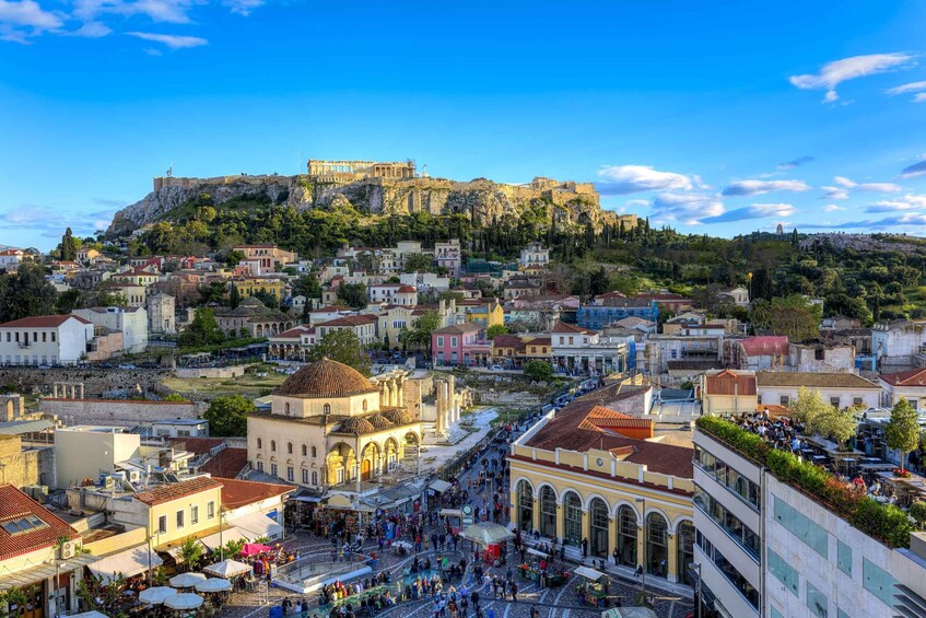 A bustling city street with historic buildings, a prominent hilltop fortress, and a clear blue sky. 3
