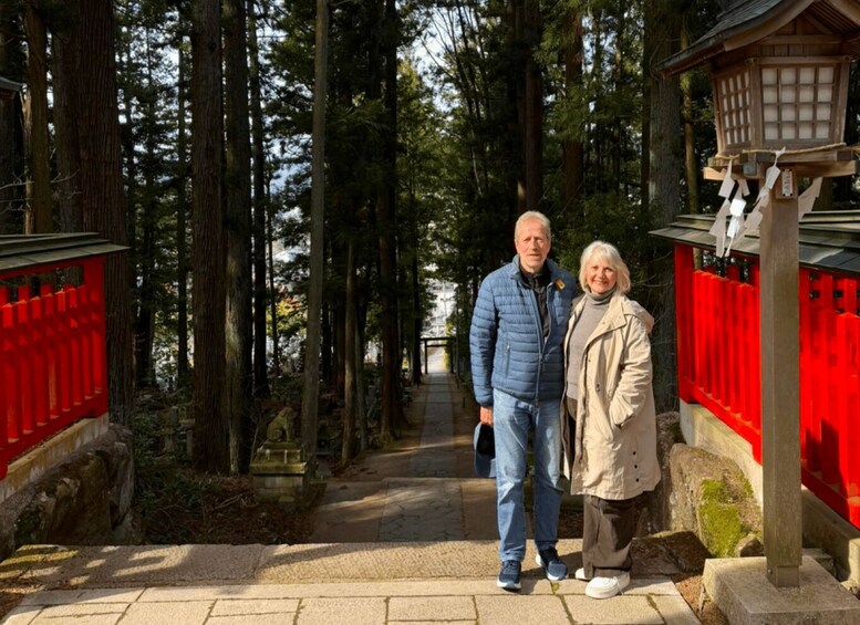 Two people standing on a stone pathway flanked by red fences and tall trees. 1