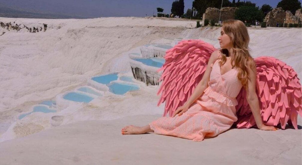 A person sitting on a white surface with pink wings, surrounded by white and blue mineral pools. 5