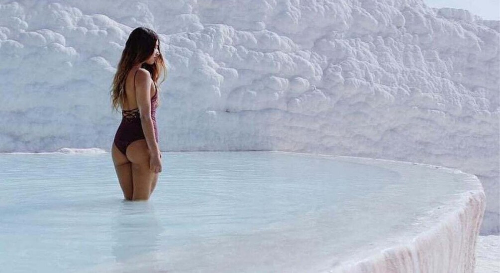 A person in a purple swimsuit standing in a clear pool with a white, textured background. 4