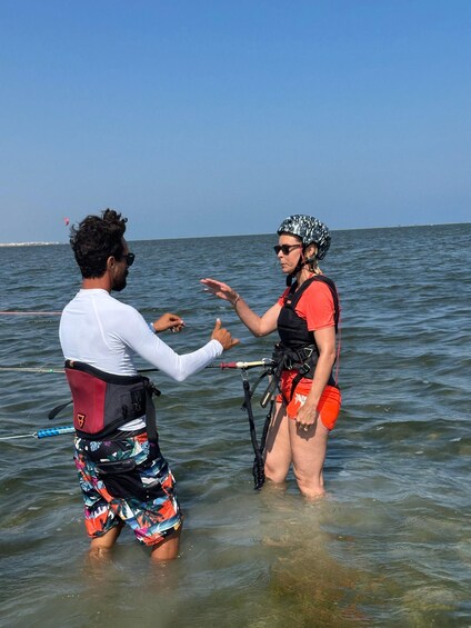 Two people in the water, one wearing a helmet and life vest, the other in a white shirt and colorful shorts, with a parasail in the background. 5