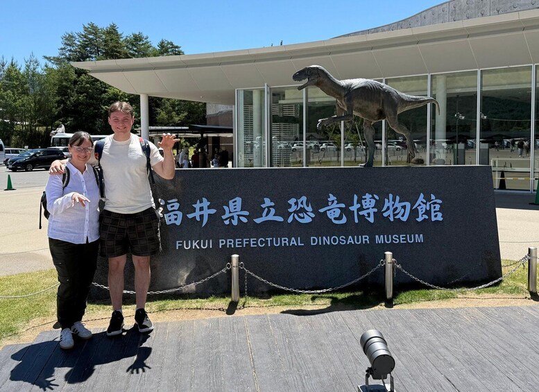 A couple posing in front of the Fukui Prefectural Dinosaur Museum with a large dinosaur statue in the background. 3