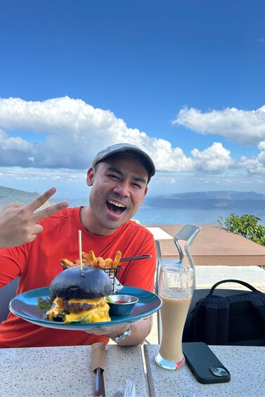 A person holding a burger with a drink and a phone on a table with a scenic view. 4