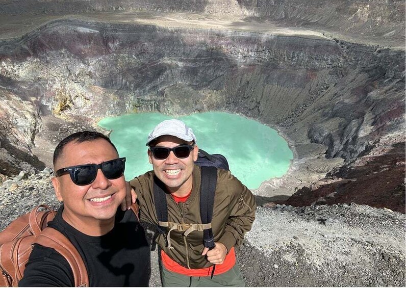 Two people taking a selfie with a large crater and turquoise water in the background. 1