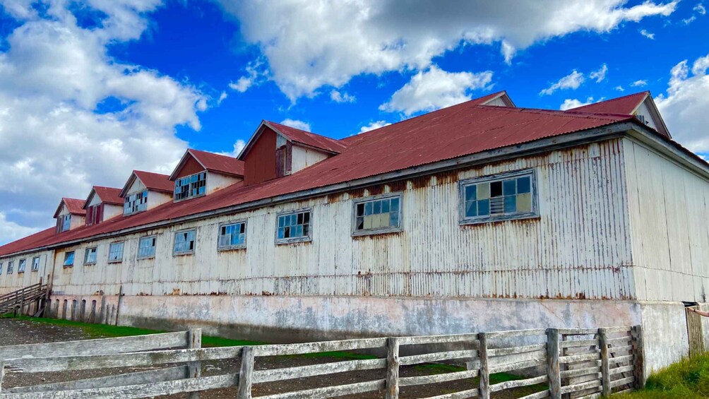 A large, weathered wooden building with a red roof, surrounded by a wooden fence. 2
