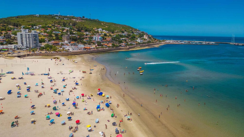 A crowded beach with people sunbathing and swimming, a clear blue sea, and a hill with buildings in the background. 1