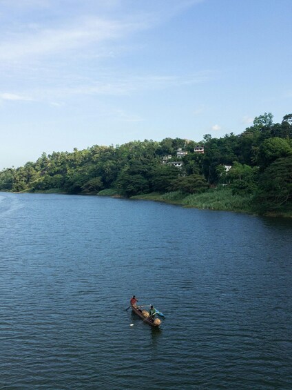 A calm river with a small boat carrying two people, surrounded by dense greenery and a few buildings on the opposite bank. 2