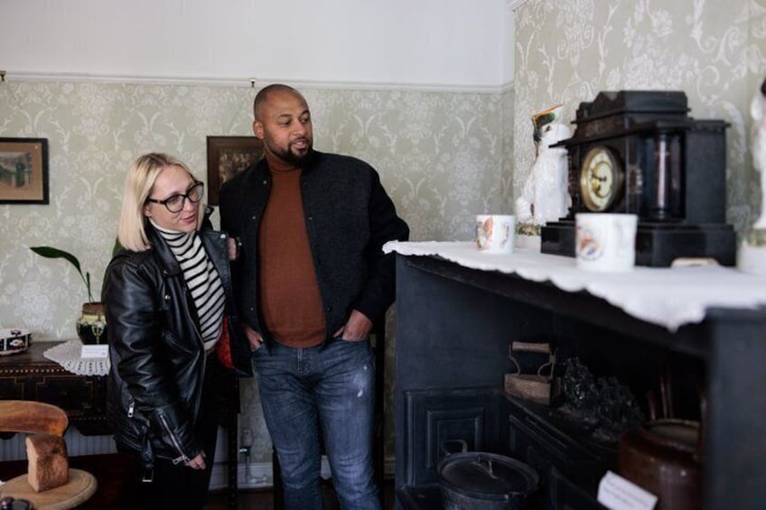 Two people standing in a room with vintage decor, including a clock and a shelf with jars. 5