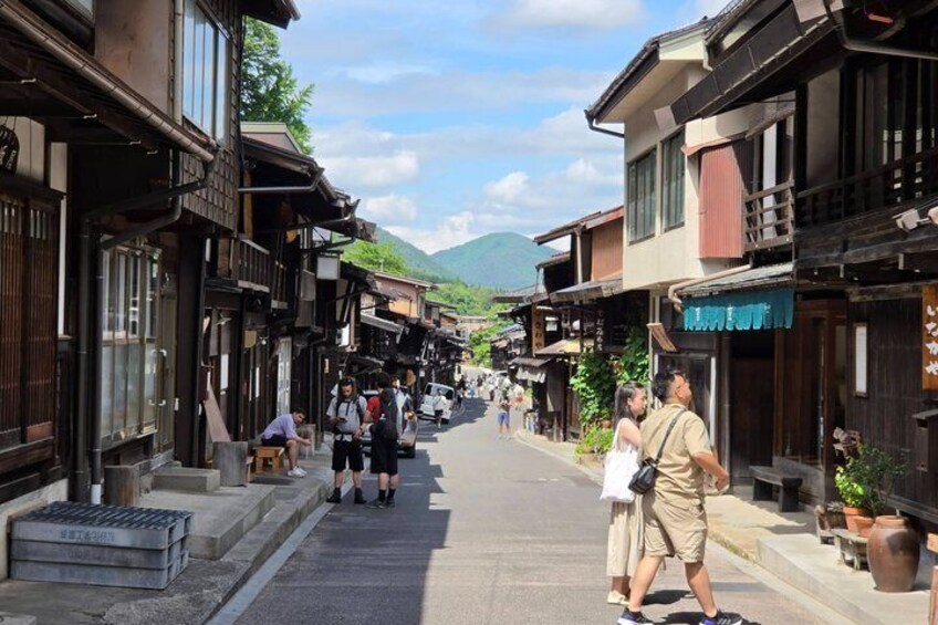 A narrow street lined with traditional wooden buildings, a few pedestrians, and a mountain in the background. 3