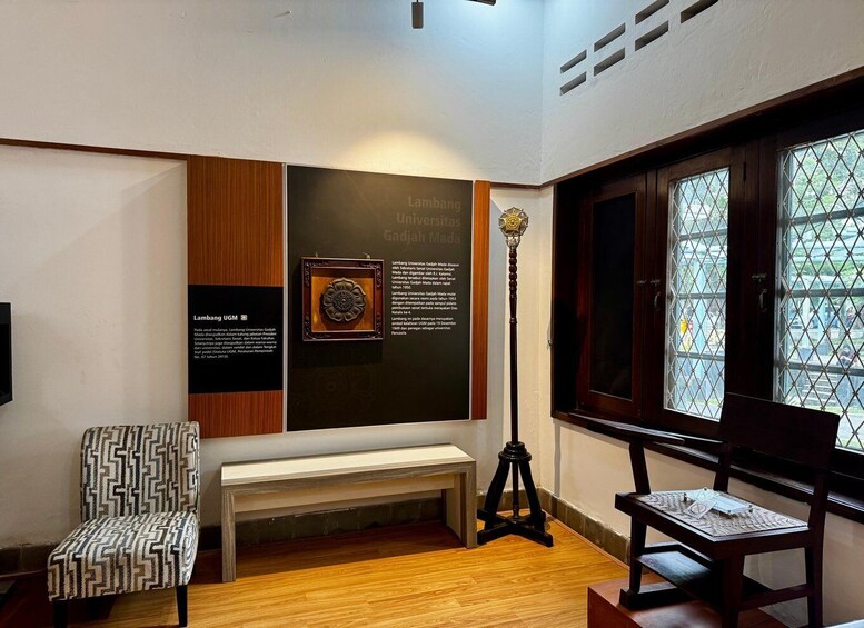 A room with a patterned chair, a bench, a table, a floor lamp, and a wall with informational plaques. 4