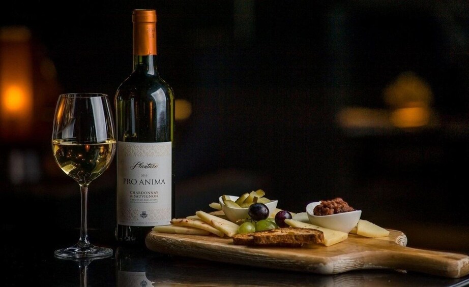 A bottle of Pro Anima wine, a glass of white wine, and a wooden board with cheese, grapes, and olives. 1