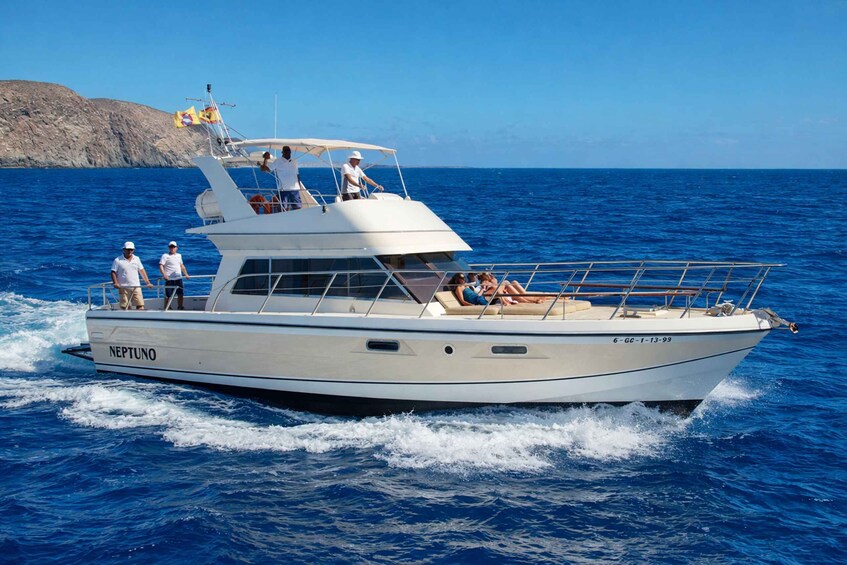 A white yacht named 'NEPTUNO' with passengers on deck, sailing on blue waters with a rocky coastline in the background. 1
