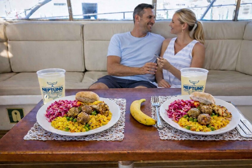 Two people seated on a couch with a table between them, each with a plate of food and a drink labeled 'NEPTUNO.' 3