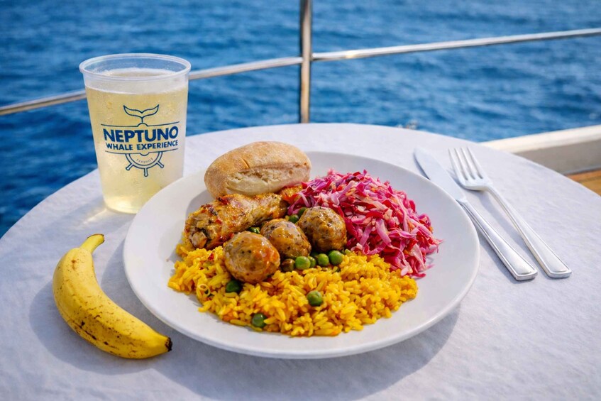 A plate of food with rice, peas, meatballs, and red cabbage, a banana, and a drink labeled 'Neptuno Whale Experience' on a boat. 5
