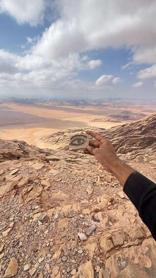 A hand holding a small stone with an eye carved into it, overlooking a desert landscape with rocky terrain and distant mountains under a partly cloudy sky. 3