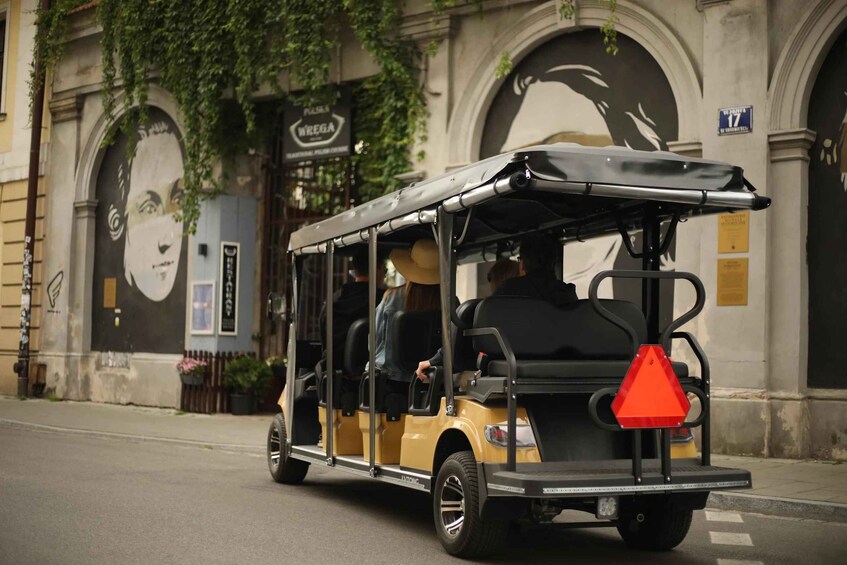 A yellow trolley with a black roof and a red triangular sign on the back, parked on a street with buildings and a mural in the background. 4