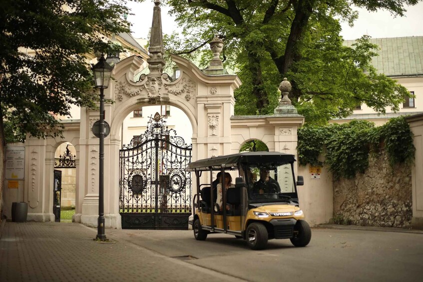 A yellow golf cart parked in front of an ornate gate with a wrought iron design, a street lamp, and a building with classical architecture. 1