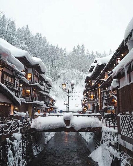 A snow-covered street with buildings on either side, a canal in the center, and a snowy forest in the background. 3