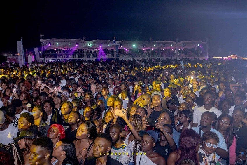 A large outdoor gathering with people wearing yellow face paint, a stage with purple lighting, and a crowd of spectators. 1