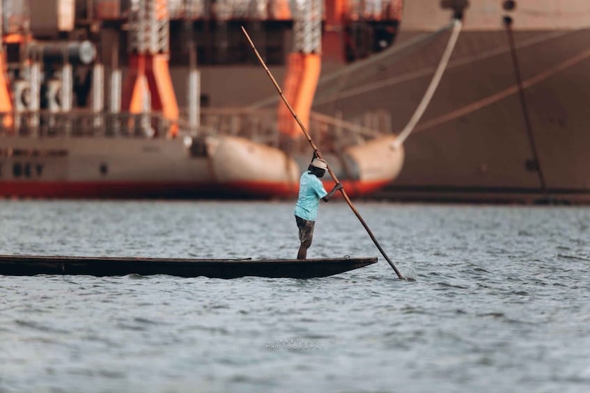 A person standing on a narrow boat in the water, holding a long pole, with a large ship in the background. 2