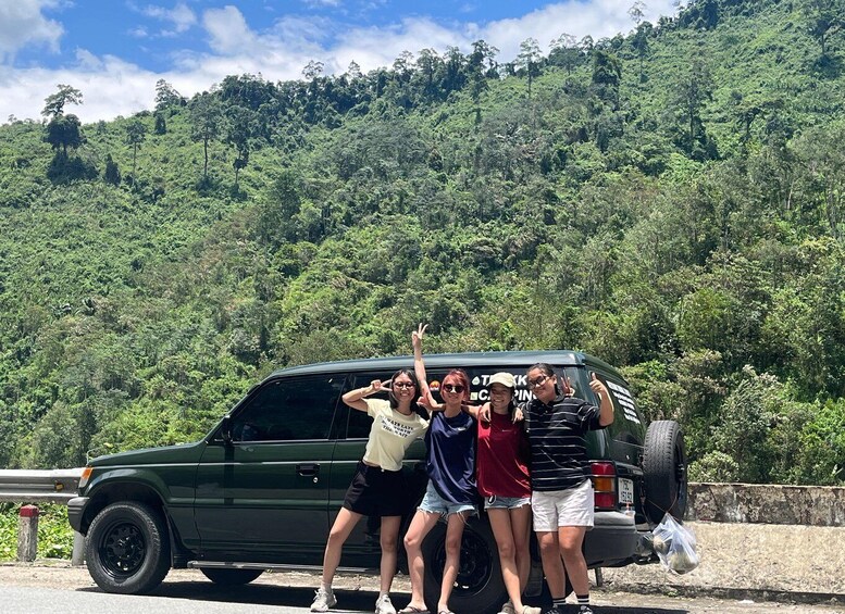 A group of people posing in front of a green SUV with a forested hillside in the background. 5
