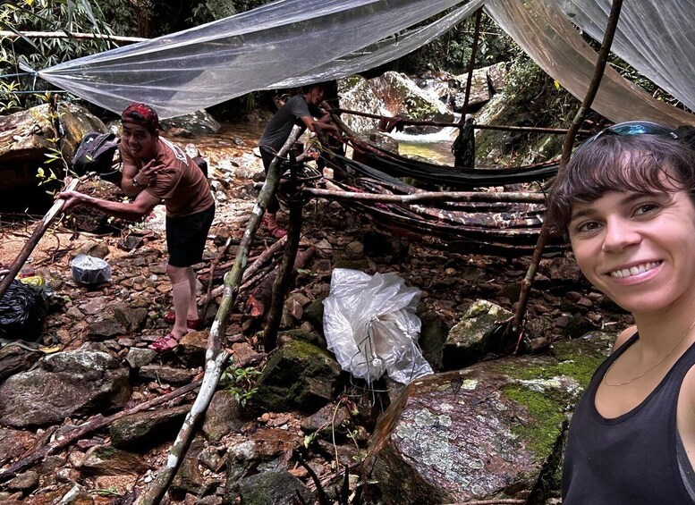 A person standing in a rocky outdoor area with a makeshift shelter and another person working on a structure. 1