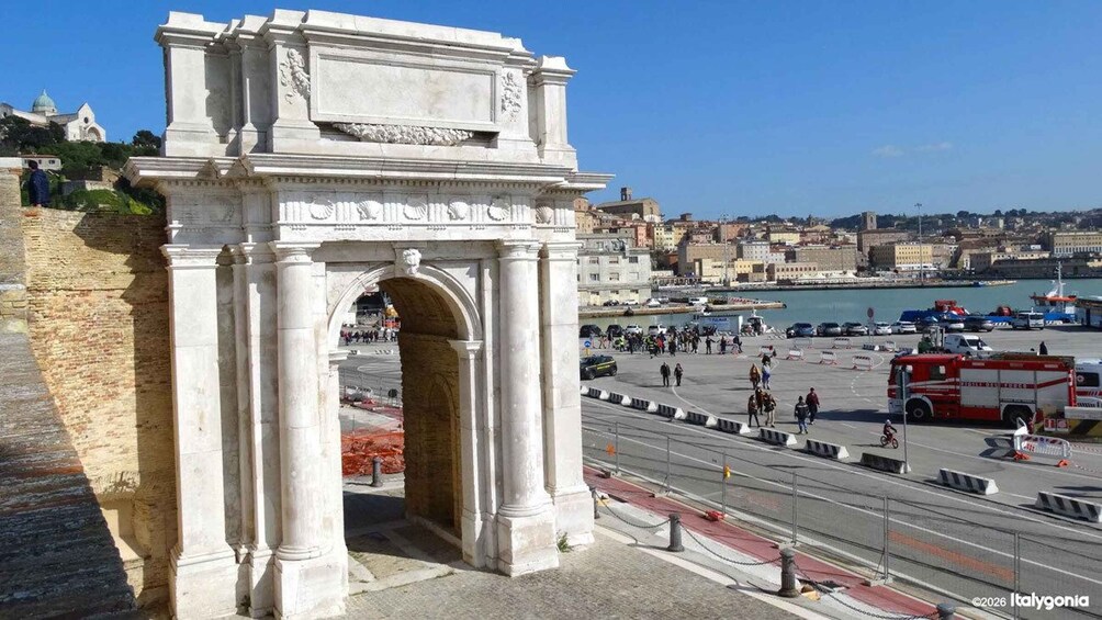 An ancient archway with classical columns, overlooking a busy harbor with boats and a red bus. 4